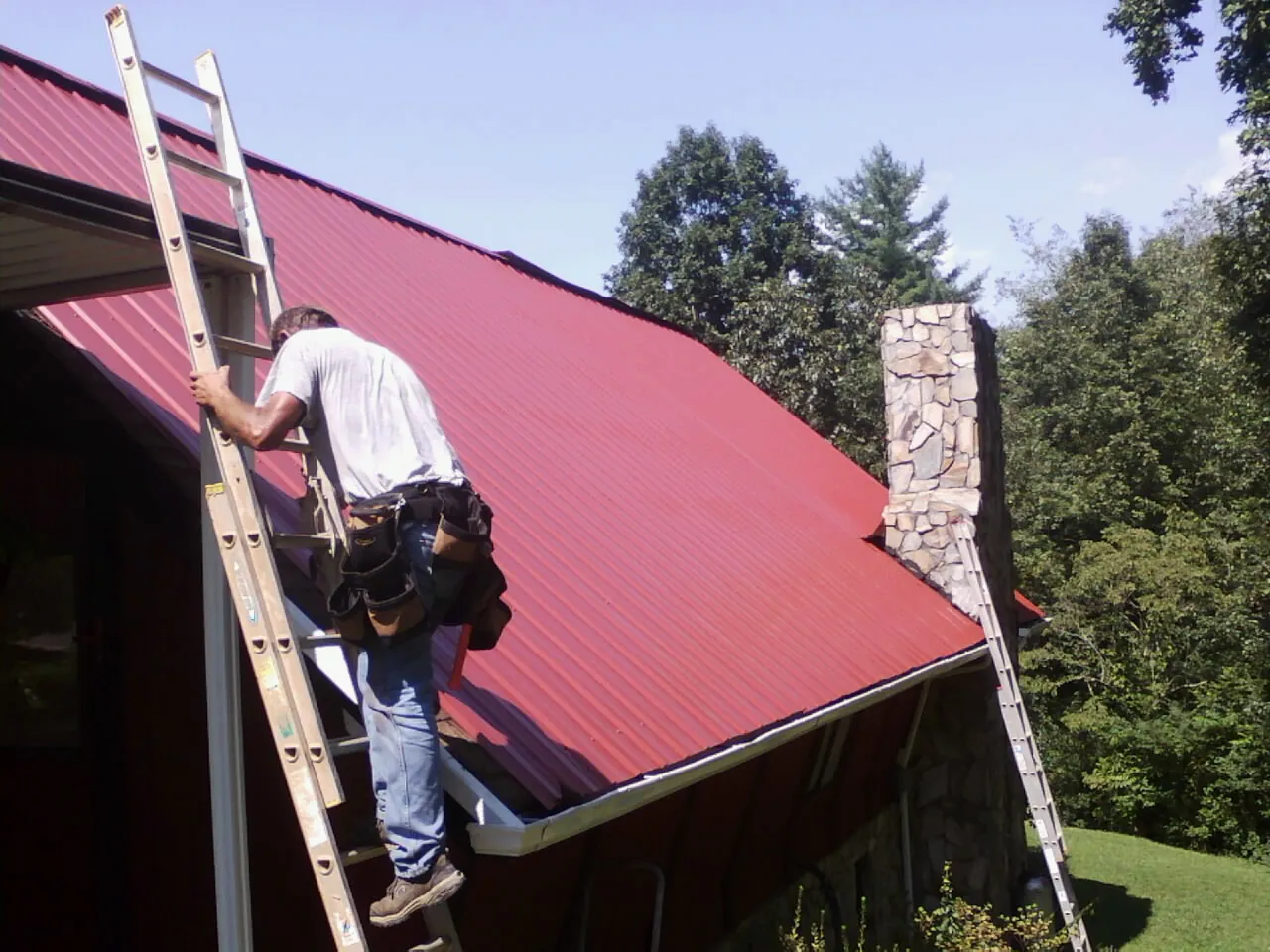 Roofer on a ladder beside a red metal roof and stone chimney — Soffit & Fascia Repair by Roofing Brothers in Onancock